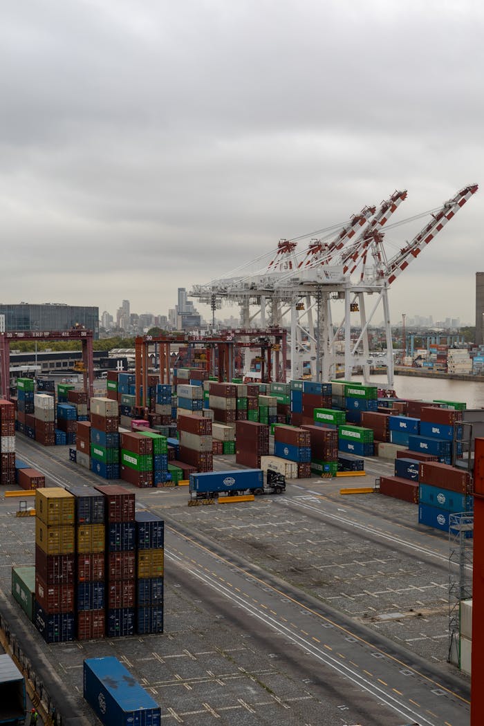 Overhead shot of a bustling container port with cranes and stacked shipping containers under a cloudy sky.